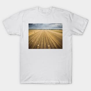 Aerial view of round hay bales on stubble with a village in the background T-Shirt